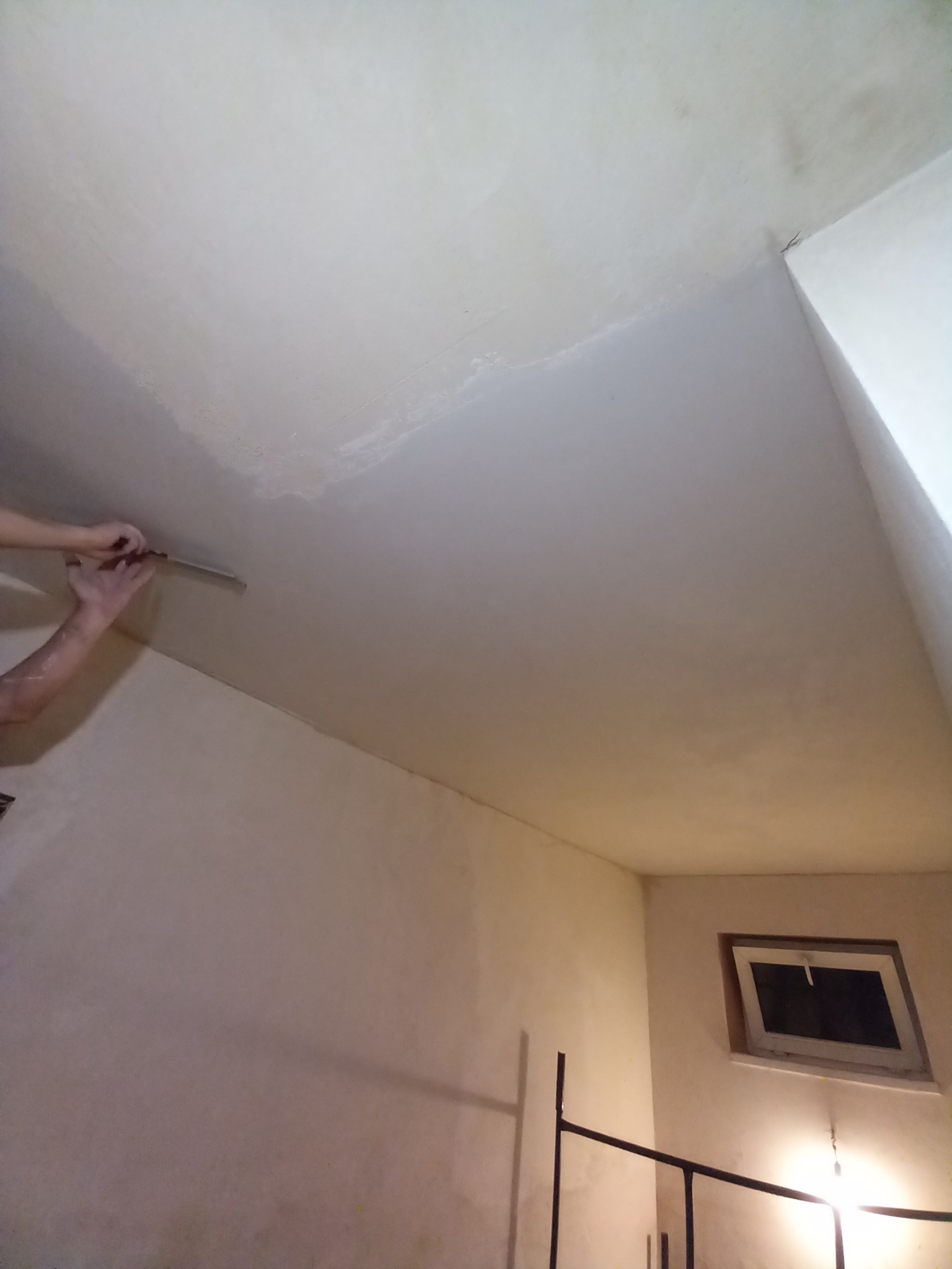 Nakładanie warstwy wykończeniowej na sufit z użyciem szpachelki, widoczne niedoskonałości powierzchni i fragment okna w tle.