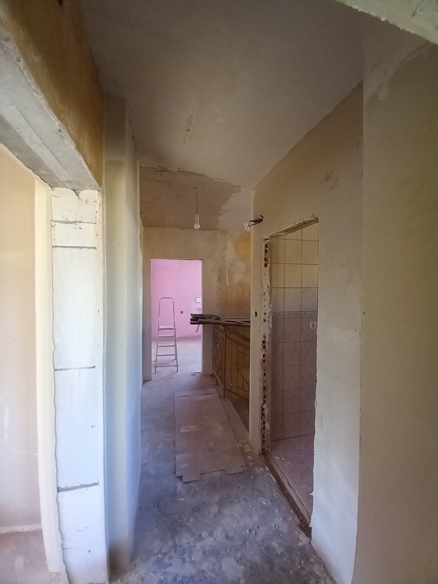 Wnętrze w trakcie remontu: surowe ściany z widocznymi śladami prac, fragment łazienki z kafelkami, drabina w tle, podłoga z resztkami materiałów budowlanych.