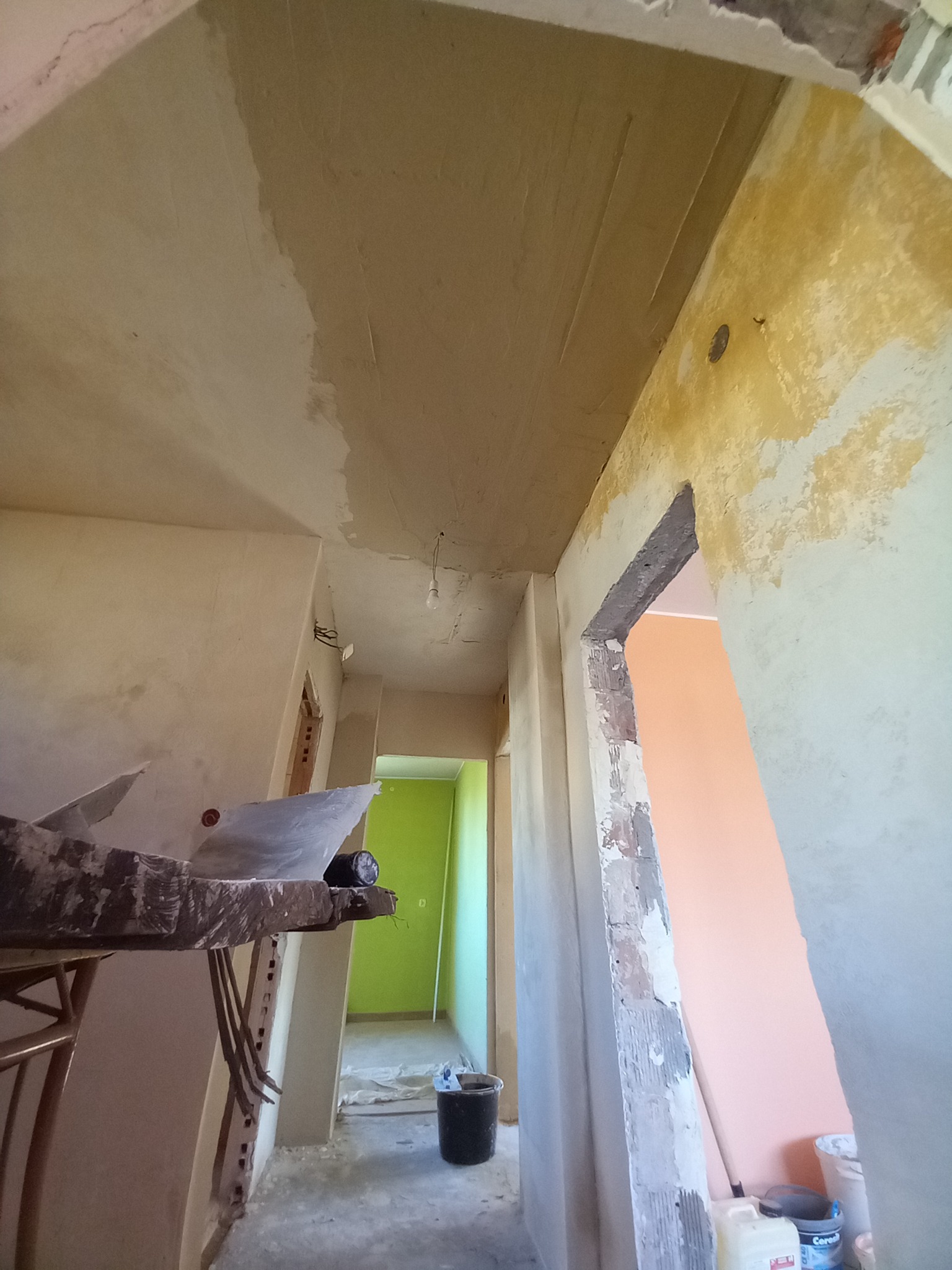 Wnętrze w trakcie remontu: częściowo wyszpachlowany sufit, fragment ściany z pozostałościami starej farby, widoczne przygotowania do dalszych prac wykończeniowych, drabina, wiadro i przejścia...