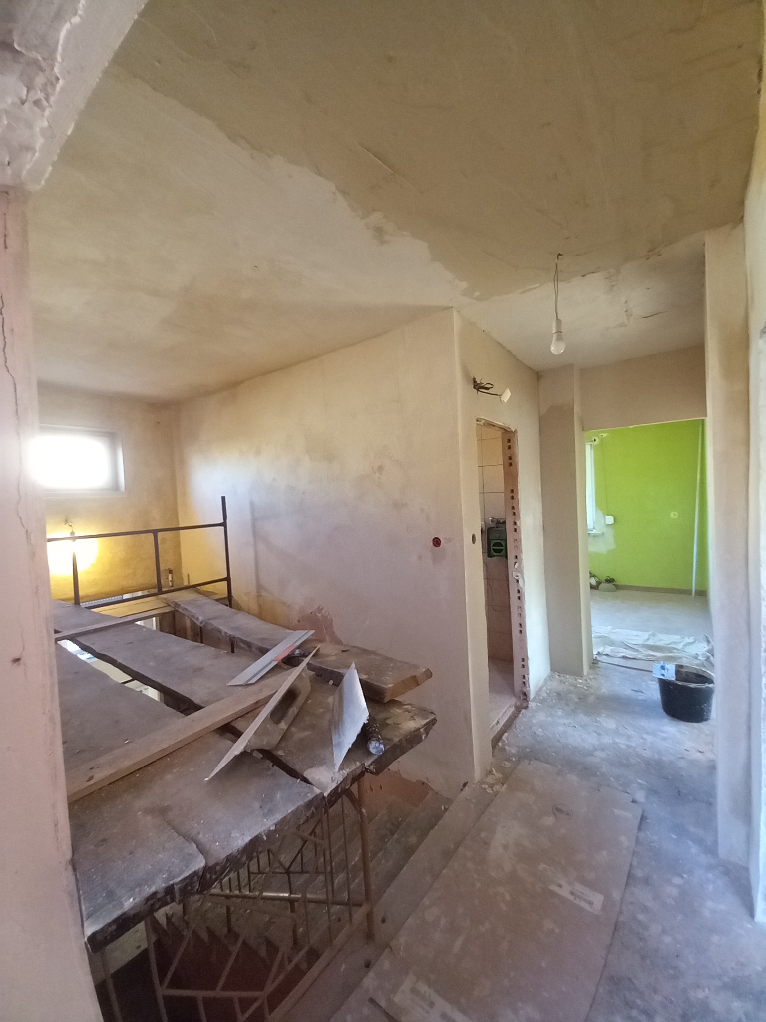 Wnętrze w trakcie prac wykończeniowych: ściany po nałożeniu gładzi, widoczne rusztowanie, narzędzia i surowa żarówka zwisająca z sufitu, w tle widoczne kolejne pomieszczenie z zieloną ścianą.