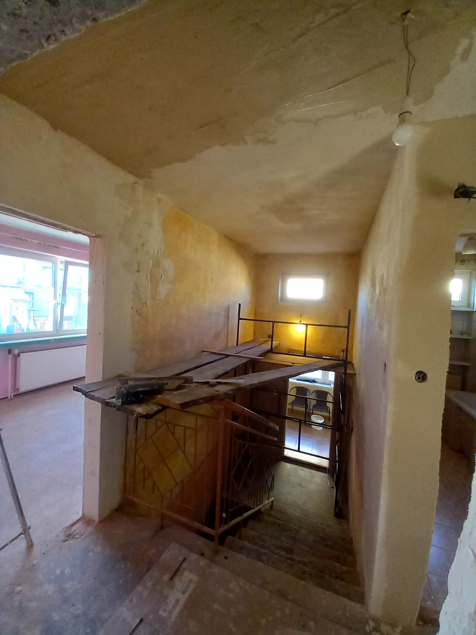 Klatka schodowa w trakcie remontu: surowe ściany z widocznymi śladami po wcześniejszych pracach, zabezpieczone prowizoryczne barierki i schody w stanie wymagającym odnowienia, z widocznym...