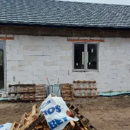 Surowy stan budynku z trzema oknami i drzwiami balkonowymi, widoczne palety i materiały budowlane na zewnątrz, dach pokryty dachówką. Budowa w trakcie.