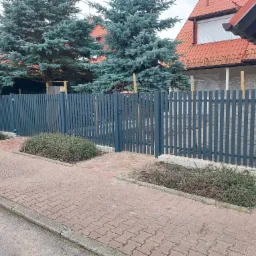 Nowoczesne, szare ogrodzenie z pionowych sztachet z furtką, widoczne z poziomu chodnika z kostki brukowej, z przodu rabaty z niską roślinnością, w tle dom z czerwoną dachówką i świerki.
