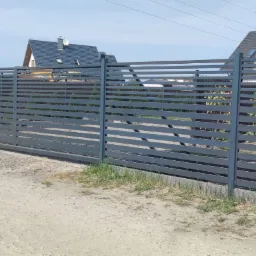 Nowoczesna, pozioma brama przesuwna i ogrodzenie z metalowych paneli w kolorze grafitowym na tle domów jednorodzinnych w słoneczny dzień.