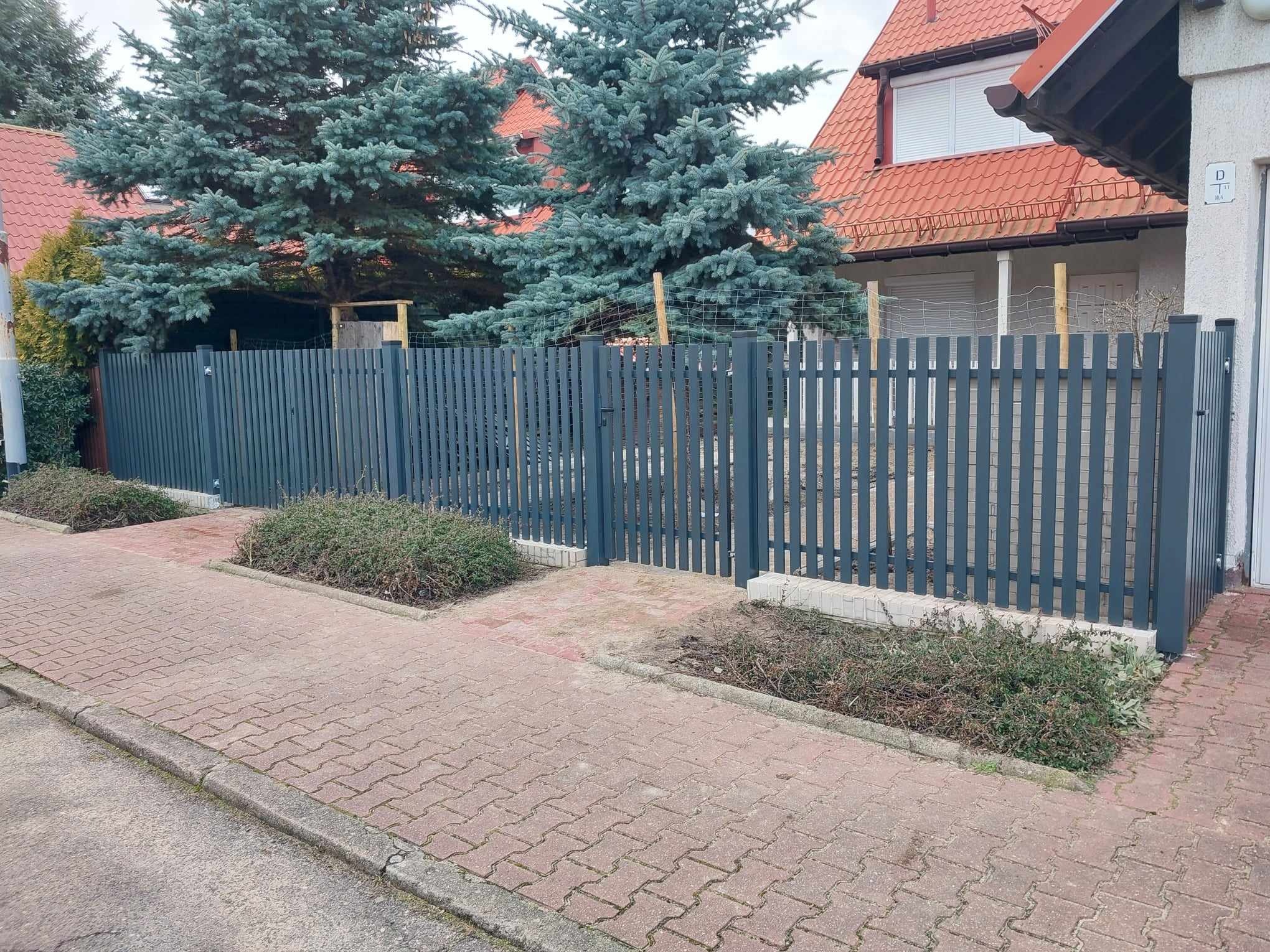 Nowoczesne, szare ogrodzenie z pionowych sztachet z furtką, widoczne z poziomu chodnika z kostki brukowej, z przodu rabaty z niską roślinnością, w tle dom z czerwoną dachówką i świerki.