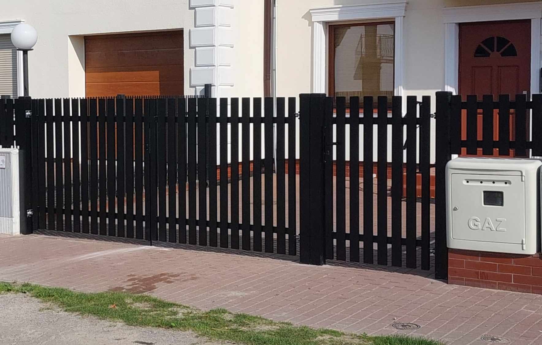 Czarna, pionowa brama z drewnianych sztachet, z widoczną skrzynką gazową po prawej stronie, na tle domu z brązowymi drzwiami garażowymi i czerwonymi drzwiami wejściowymi.
