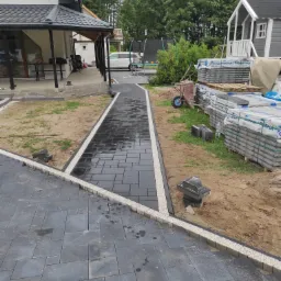 Ułożona ścieżka z ciemnoszarej kostki brukowej z białym obramowaniem z drobnych kamyczków, prowadząca od domu do ogrodu, z paletami kostki w tle i taczką obok.