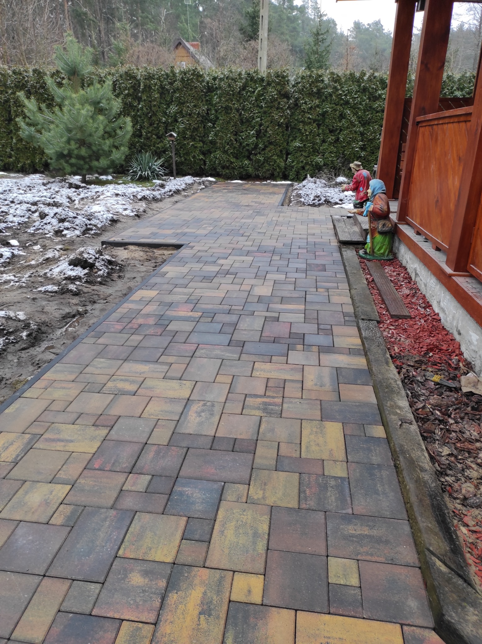 Ułożona ścieżka z wielokolorowej kostki brukowej prowadząca wzdłuż drewnianej altany, częściowo pokryta śniegiem, z dekoracyjnymi figurkami ogrodowymi.