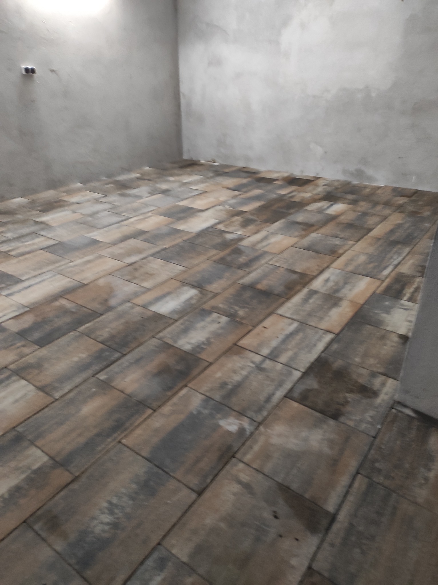 Ułożona, prostokątna kostka w odcieniach brązu i szarości na podłodze w surowym pomieszczeniu z szarą, nieotynkowaną ścianą i widocznymi puszkami elektrycznymi.