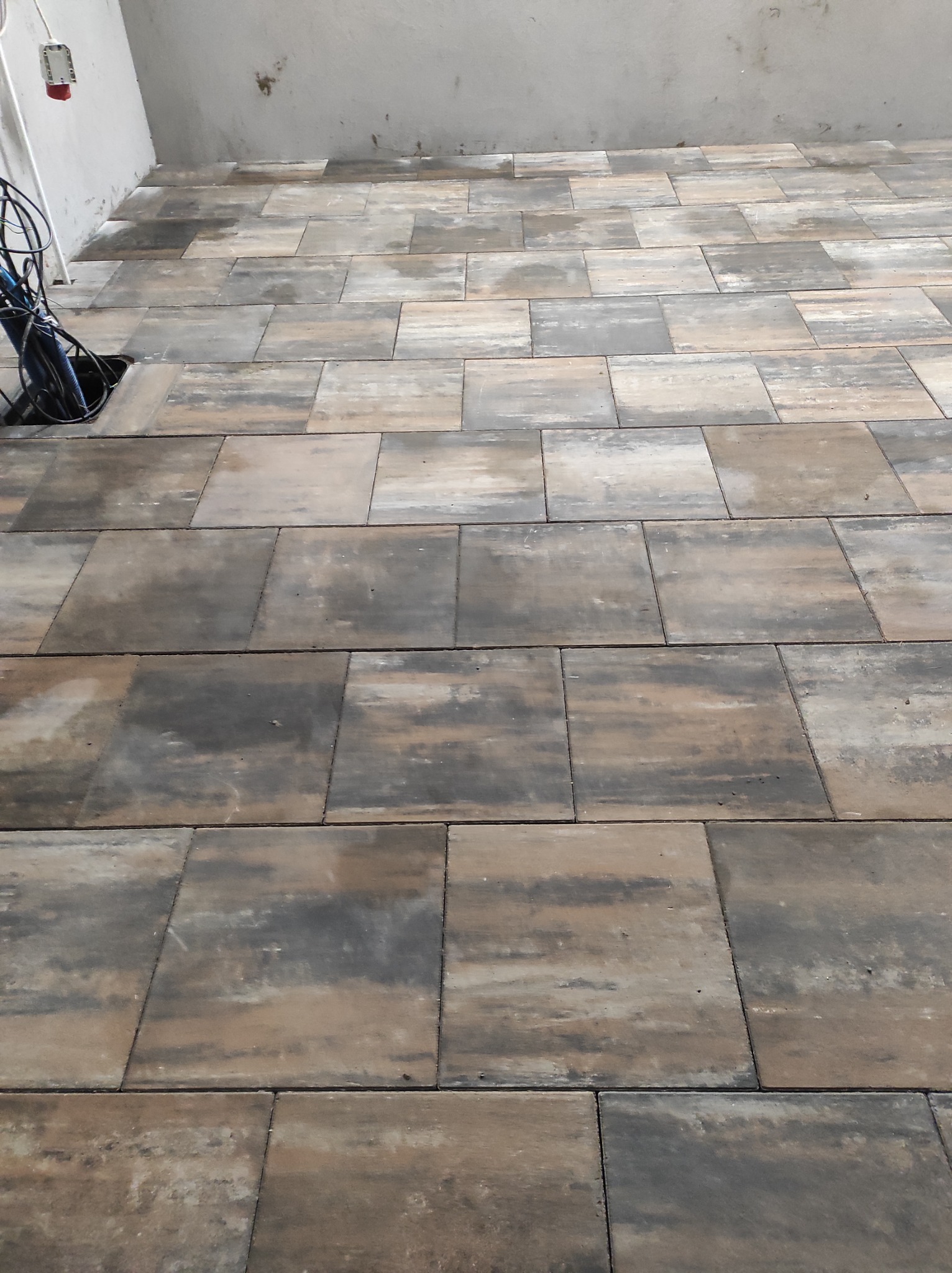 Ułożona, brązowo-szara kostka brukowa na podłożu wewnątrz budynku, z widocznymi przewodami elektrycznymi w rogu pomieszczenia.