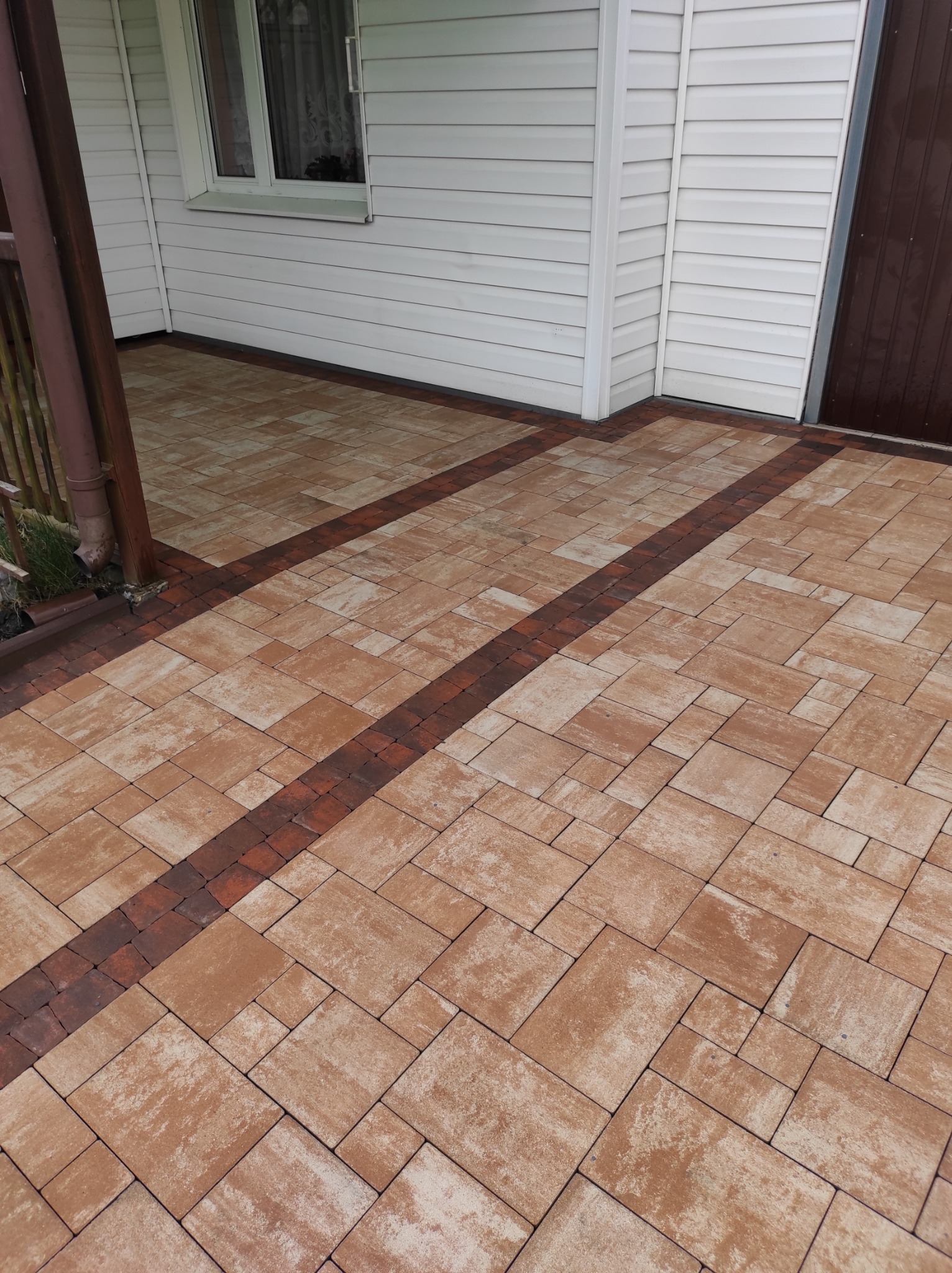 Ułożona kostka brukowa w kolorze beżowo-brązowym z ciemniejszym obramowaniem przed domem z białym sidingiem i brązowymi drzwiami.