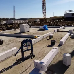Wykonywanie fundamentów z bloczków betonowych, widoczny wózek transportowy, kask ochronny i żuraw budowlany w tle.