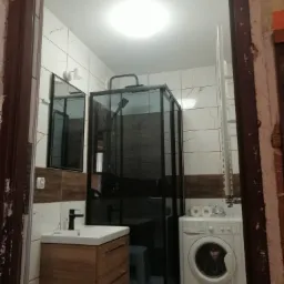 Wykończona łazienka z kabiną prysznicową typu walk-in z czarnymi ramami, umywalką z szafką w kolorze drewna, pralką i grzejnikiem drabinkowym, widok z perspektywy wejścia.