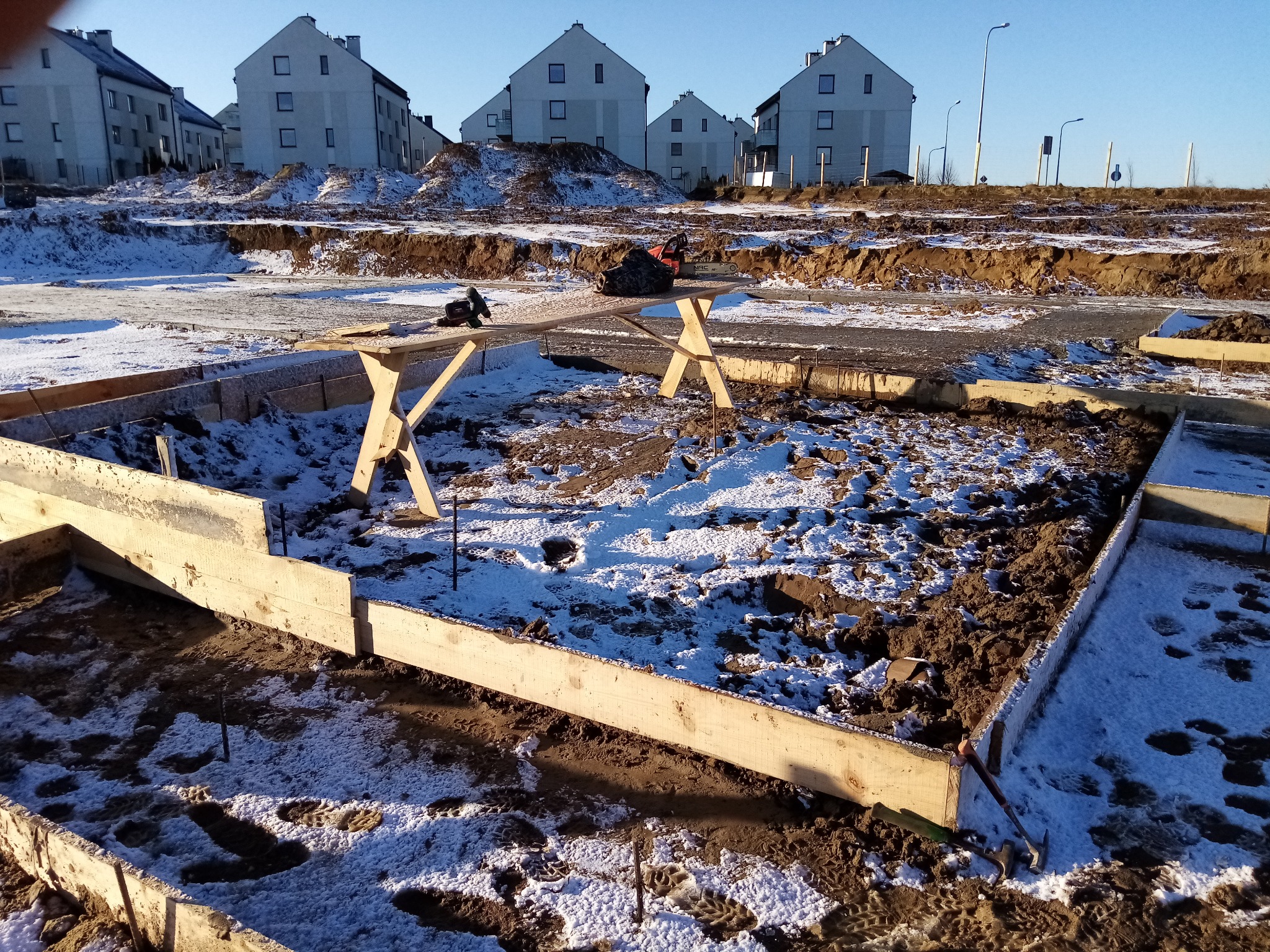 Deskowanie fundamentu z drewnianych belek na zasypanym śniegiem placu budowy w słoneczny dzień, w tle widoczne budynki mieszkalne.