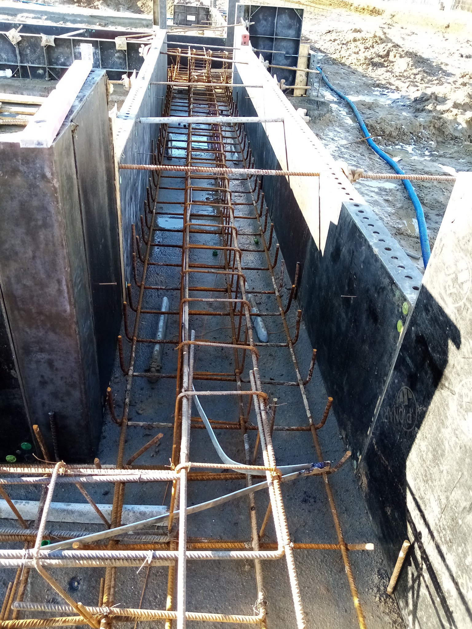 Zbrojenie fundamentu w szalunku z widocznymi elementami stalowymi, rurami instalacyjnymi i ziemnym podłożem.