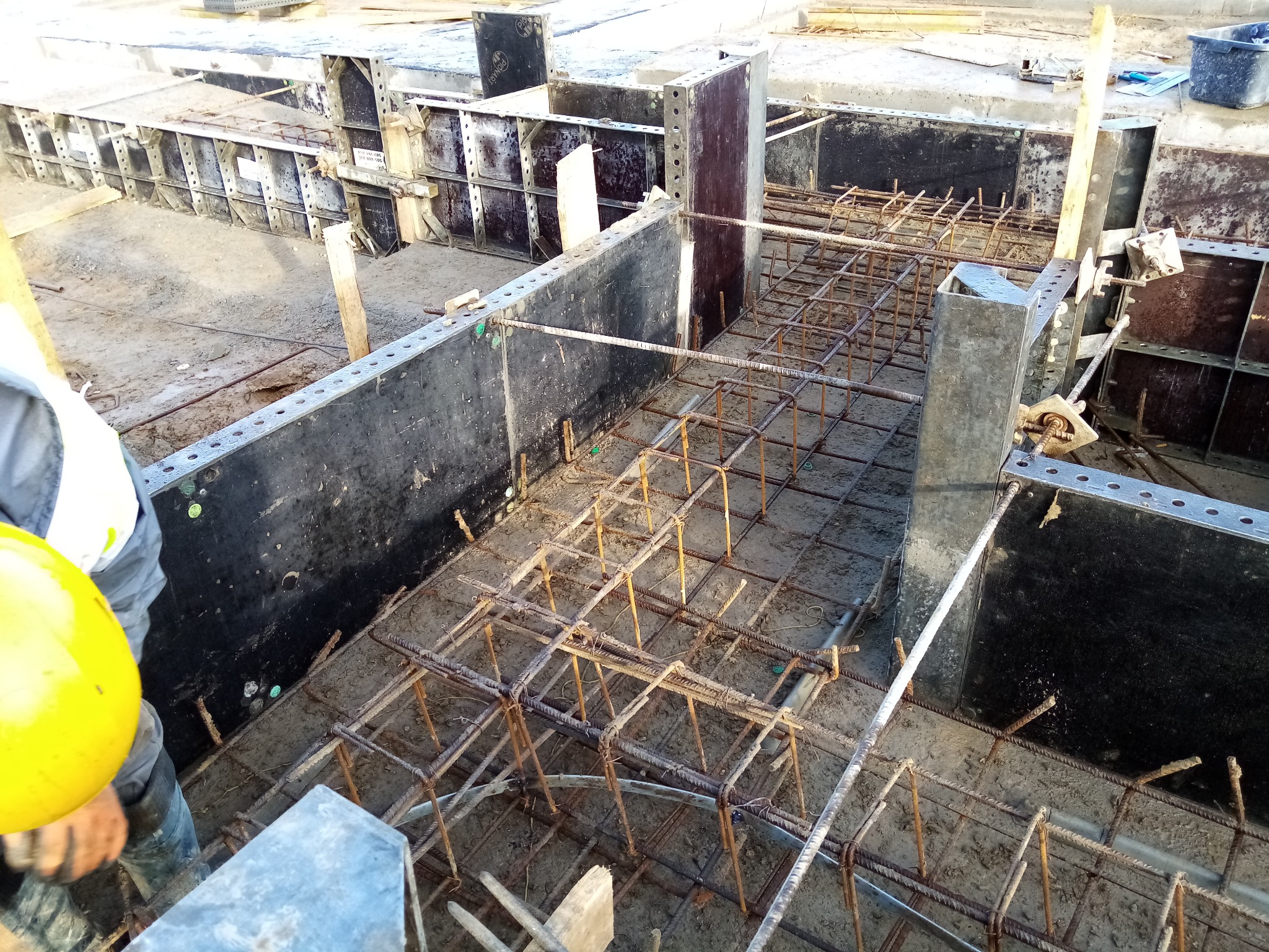 Przygotowanie zbrojenia i szalunków pod wylewkę fundamentu, widoczny fragment żółtego kasku pracownika budowlanego.