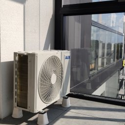 INSTALKLIMA SPÓŁKA Z OGRANICZONĄ ODPOWIEDZIALNOŚCIĄ - Biała jednostka zewnętrzna klimatyzacji AUX na balkonie nowoczesnego budynku z dużymi oknami, widoczna z boku, zamontowana na podkładkach antywibracyjnych.