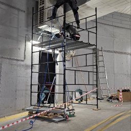 INSTALKLIMA SPÓŁKA Z OGRANICZONĄ ODPOWIEDZIALNOŚCIĄ - Pracownik w kombinezonie montuje instalację wentylacyjną na wysokości, stojąc na rusztowaniu obok jednostki klimatyzacji w szarym pomieszczeniu.