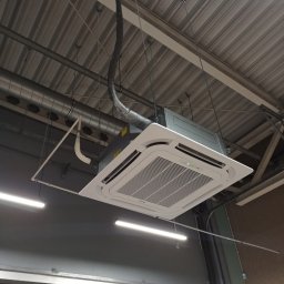INSTALKLIMA SPÓŁKA Z OGRANICZONĄ ODPOWIEDZIALNOŚCIĄ - Sufitowa klimatyzacja kasetonowa, zawieszona na linkach w pomieszczeniu przemysłowym. Widoczne przewody i instalacja. Oświetlenie jarzeniowe.