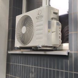 INSTALKLIMA SPÓŁKA Z OGRANICZONĄ ODPOWIEDZIALNOŚCIĄ - Biały klimatyzator marki Konsi zamontowany na parapecie okna z ciemnymi płytkami. Widoczny fragment ławki. Ujęcie z dołu.