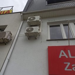 INSTALKLIMA SPÓŁKA Z OGRANICZONĄ ODPOWIEDZIALNOŚCIĄ - Dwa klimatyzatory KONSI zamontowane jeden nad drugim na szarej ścianie budynku z widoczną rynną i fragmentem czerwonego szyldu reklamowego. Pochmurne niebo w tle.