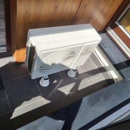 INSTALKLIMA SPÓŁKA Z OGRANICZONĄ ODPOWIEDZIALNOŚCIĄ - Kompaktowa jednostka zewnętrzna klimatyzacji marki Konsi, ustawiona na białych wspornikach, na ciemnych płytkach, w pobliżu drewnianych drzwi i okna.