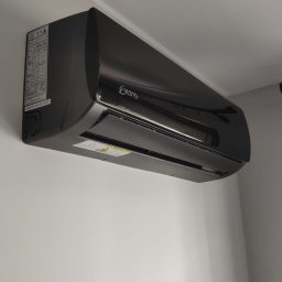 INSTALKLIMA SPÓŁKA Z OGRANICZONĄ ODPOWIEDZIALNOŚCIĄ - Czarny klimatyzator marki Konsi zamontowany na ścianie. Widoczne oznaczenia na boku urządzenia. Minimalistyczny design i gładka powierzchnia.
