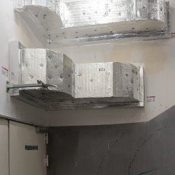 INSTALKLIMA SPÓŁKA Z OGRANICZONĄ ODPOWIEDZIALNOŚCIĄ - Dwa poziomy izolowanych kanałów wentylacyjnych, zabezpieczonych folią aluminiową, zamontowane na białej ścianie w pobliżu klatki schodowej.