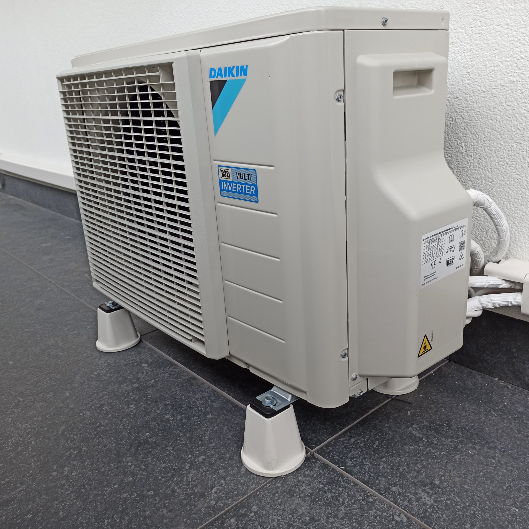 Zewnętrzna jednostka klimatyzacyjna Daikin R32 Multi Inverter zamontowana na wspornikach na ciemnej, płytkowej posadzce, przy białej ścianie.