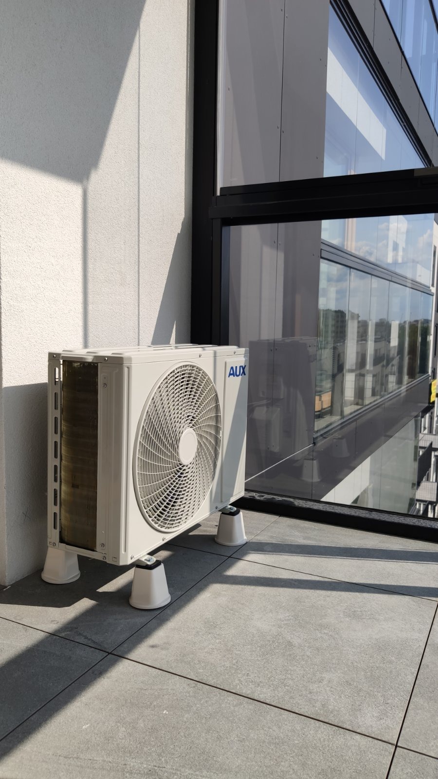 Biała jednostka zewnętrzna klimatyzacji AUX na balkonie nowoczesnego budynku z dużymi oknami, widoczna z boku, zamontowana na podkładkach antywibracyjnych.