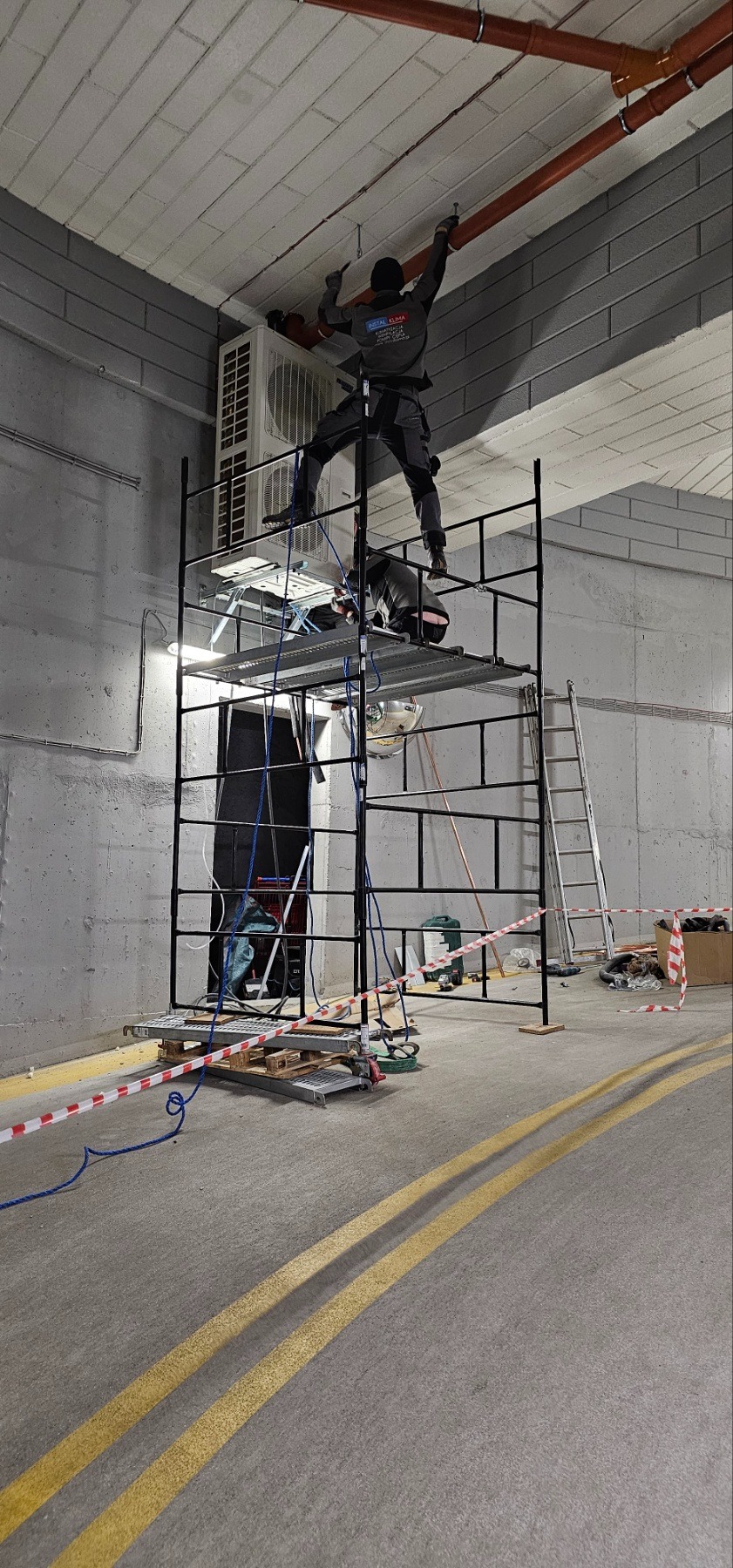 Pracownik w kombinezonie montuje instalację wentylacyjną na wysokości, stojąc na rusztowaniu obok jednostki klimatyzacji w szarym pomieszczeniu.