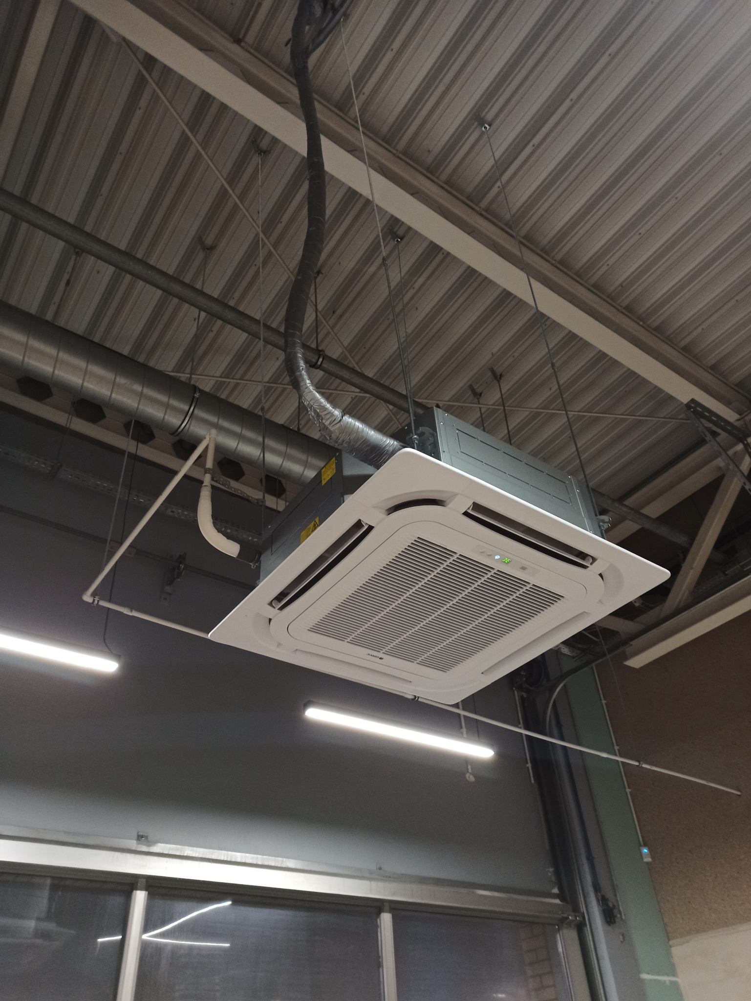 Sufitowa klimatyzacja kasetonowa, zawieszona na linkach w pomieszczeniu przemysłowym. Widoczne przewody i instalacja. Oświetlenie jarzeniowe.