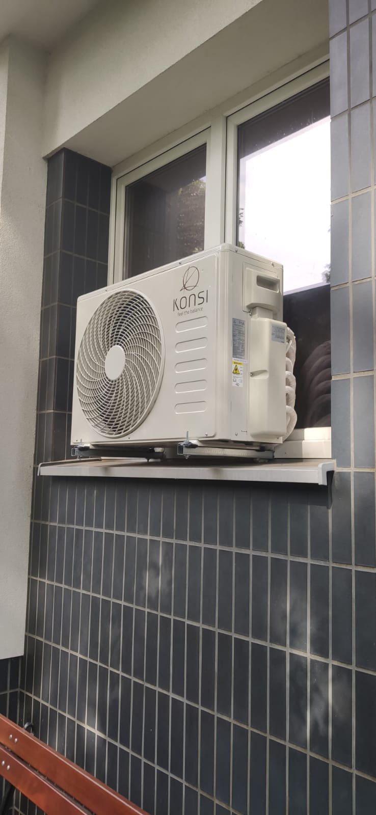 Biały klimatyzator marki Konsi zamontowany na parapecie okna z ciemnymi płytkami. Widoczny fragment ławki. Ujęcie z dołu.