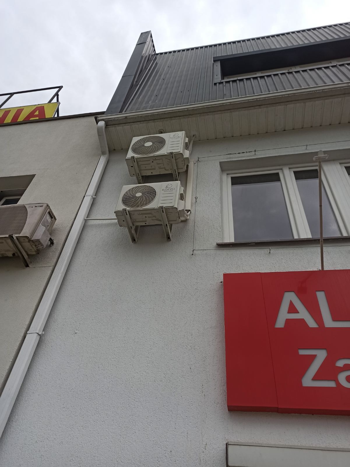Dwa klimatyzatory KONSI zamontowane jeden nad drugim na szarej ścianie budynku z widoczną rynną i fragmentem czerwonego szyldu reklamowego. Pochmurne niebo w tle.