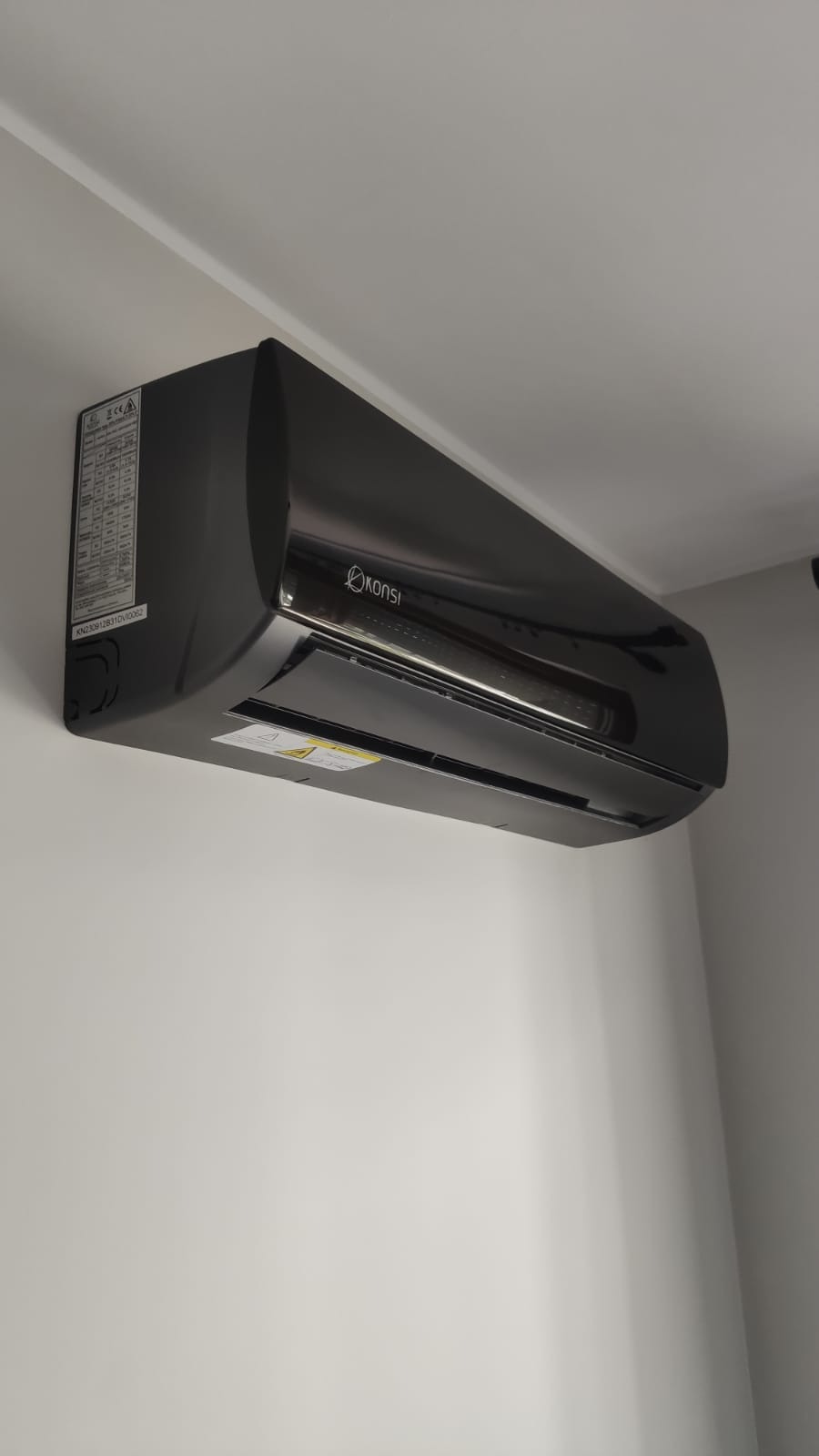 Czarny klimatyzator marki Konsi zamontowany na ścianie. Widoczne oznaczenia na boku urządzenia. Minimalistyczny design i gładka powierzchnia.