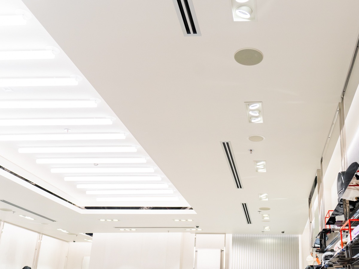 Biały sufit w sklepie z zamontowaną wentylacją mechaniczną. Widoczne kratki wentylacyjne, oświetlenie i głośniki sufitowe. Minimalistyczny design.