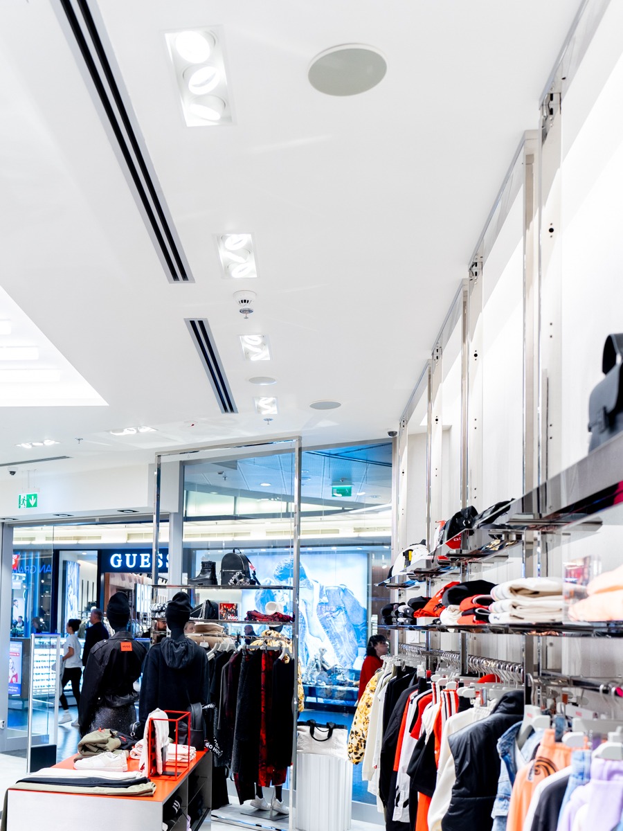 Wnętrze sklepu odzieżowego z widocznymi elementami wentylacji liniowej w suficie podwieszanym oraz okrągłymi nawiewnikami sufitowymi.