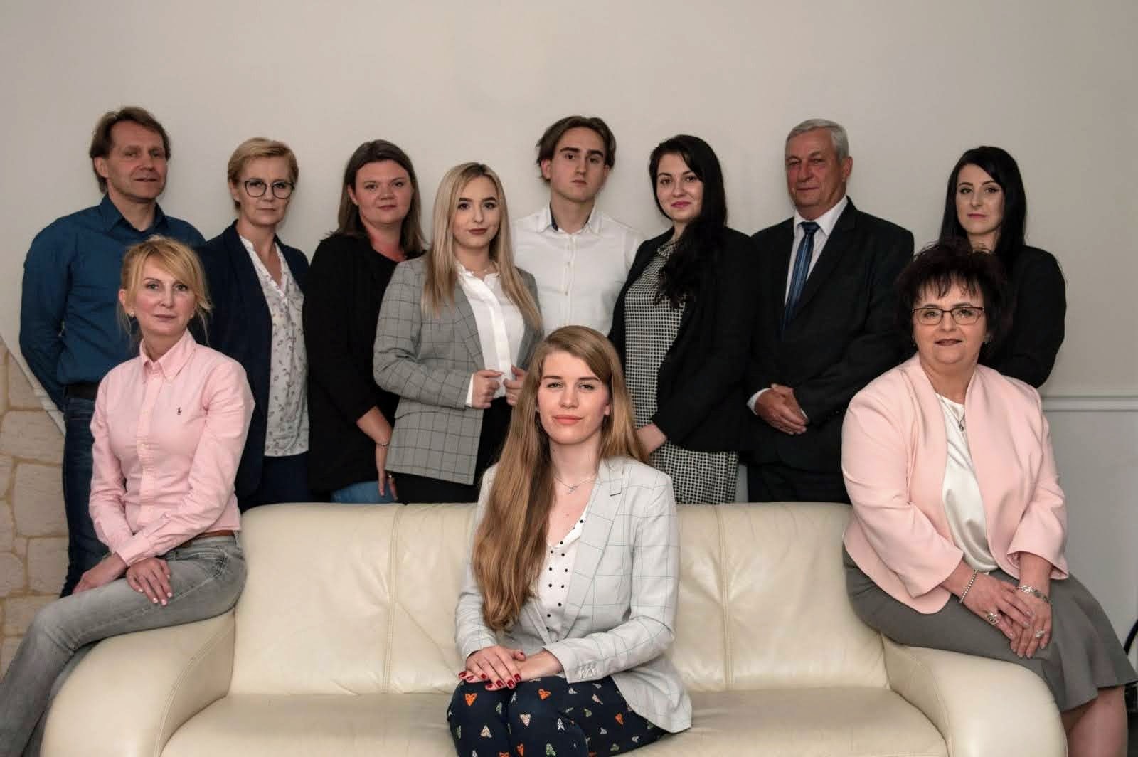 Zespół pracowników firmy ubezpieczeniowej pozuje do zdjęcia grupowego w biurze, kobiety w marynarkach i mężczyźni w koszulach i garniturach.