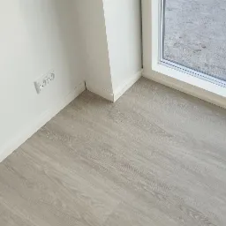 Jasnoszare panele podłogowe z widocznym usłojeniem, białe listwy przypodłogowe, gniazdko elektryczne przy białej ścianie, okno z widokiem na zewnątrz.