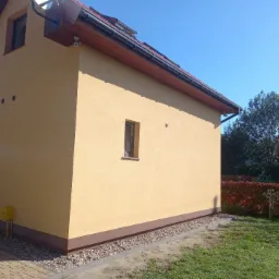 Żółta elewacja domu jednorodzinnego z brązowymi detalami wykończeniowymi, widok z boku na tle zieleni i błękitnego nieba w słoneczny dzień.