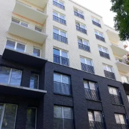 Nowoczesna elewacja budynku z ciemnej cegły na dole i jasnej u góry. Okna zabezpieczone folią. Podnośnik koszowy z pracownikiem przy balkonie.