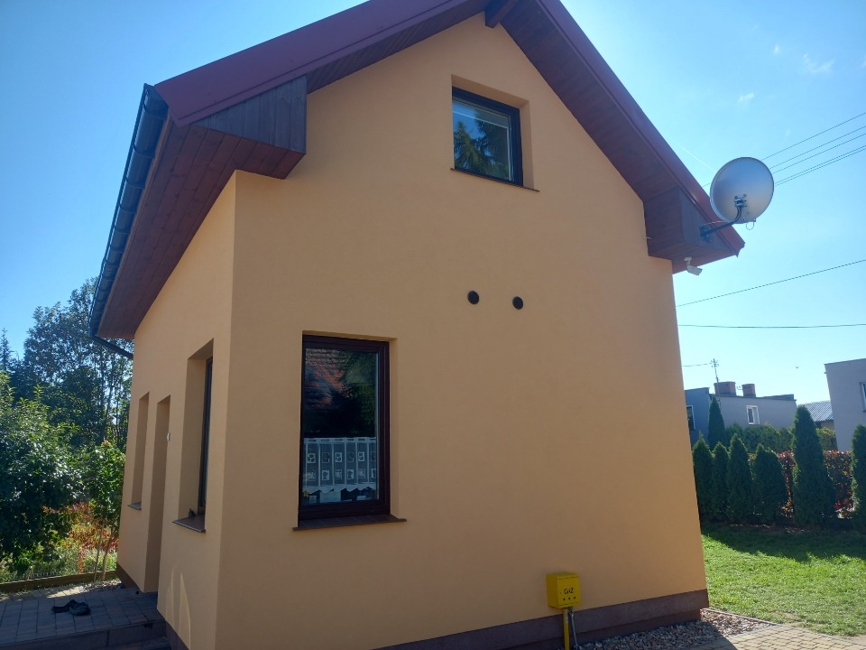 Beżowa elewacja domu jednorodzinnego z brązowym dachem, widoczna antena satelitarna, okna z ciemnymi ramami i skrzynka gazowa przy ścianie.