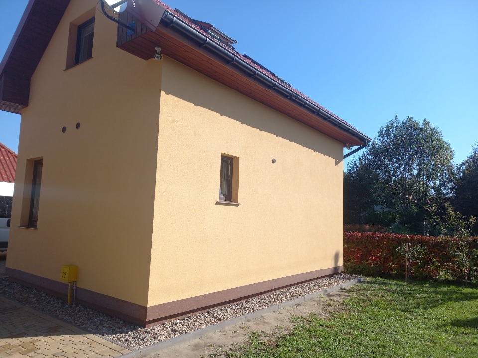 Żółta elewacja domu jednorodzinnego z brązowymi detalami wykończeniowymi, widok z boku na tle zieleni i błękitnego nieba w słoneczny dzień.