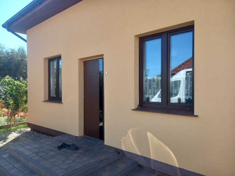 Ocieplony dom z beżową elewacją, brązowymi oknami i drzwiami. Na schodach leżą klapki, a w oknie odbija się biały samochód dostawczy.