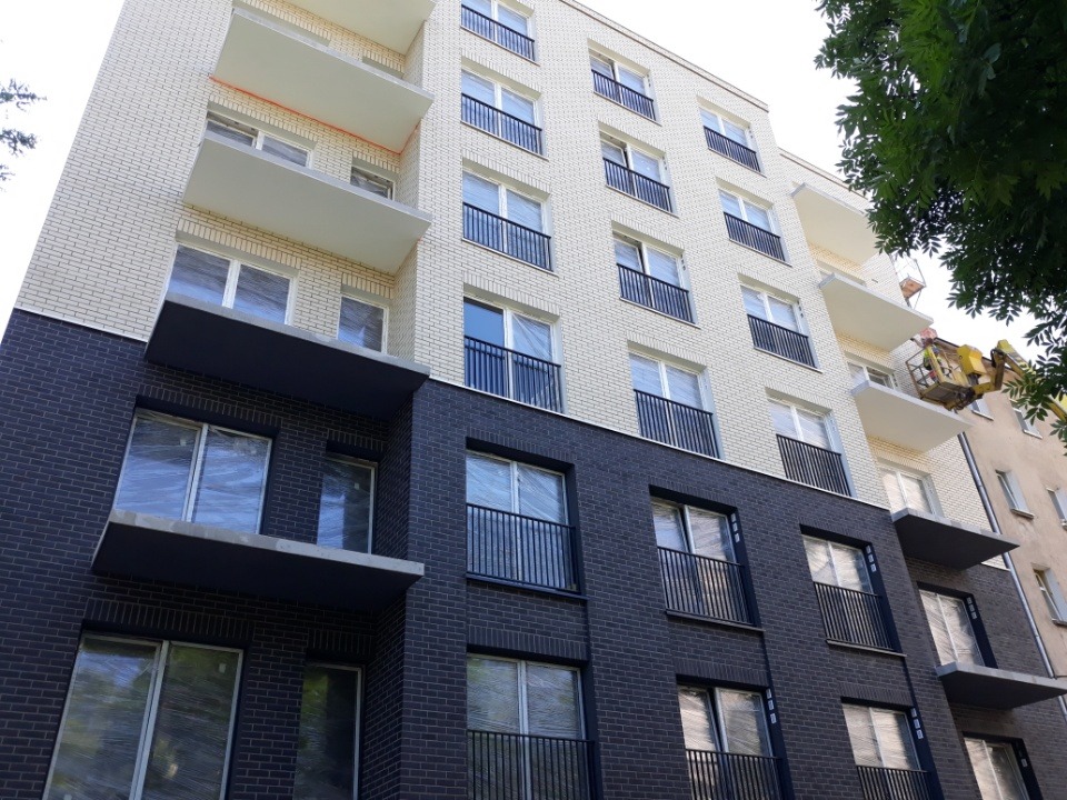 Nowoczesna elewacja budynku z ciemnej cegły na dole i jasnej u góry. Okna zabezpieczone folią. Podnośnik koszowy z pracownikiem przy balkonie.