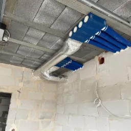 Niedokończony montaż systemu rur wentylacyjnych w surowym wnętrzu, widoczne niebieskie rury w białej piance montażowej, srebrny kanał wentylacyjny i betonowe ściany w trakcie budowy.