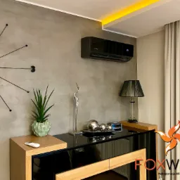 Nowoczesny czarny klimatyzator na szarej ścianie nad lakierowaną komodą w salonie z zegarem słonecznym i dekoracjami.