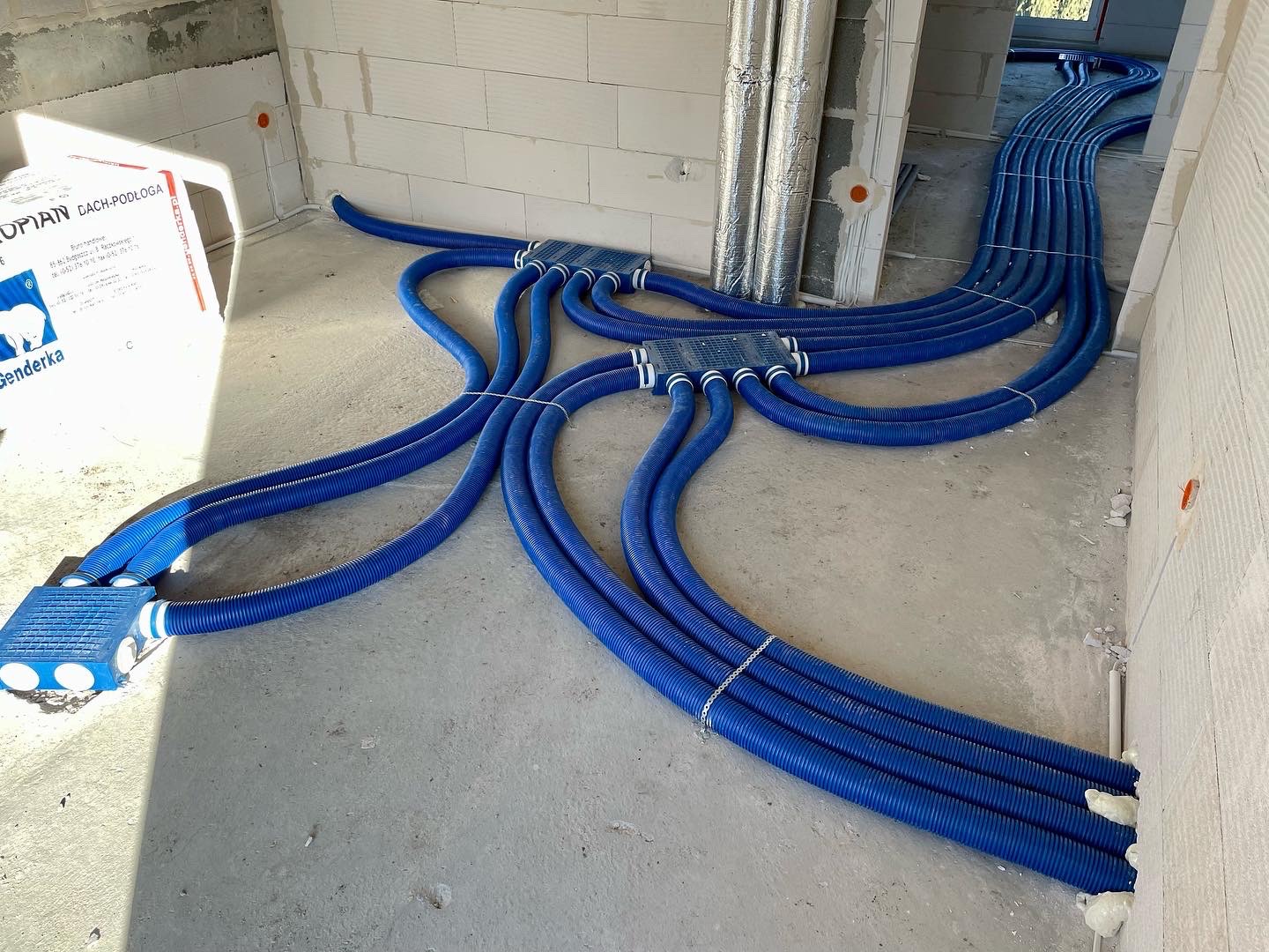 Niebieskie rury wentylacyjne ułożone na surowym betonowym podłożu w trakcie instalacji w budynku, widoczne skrzynki rozdzielcze i izolacja termiczna rur w tle.