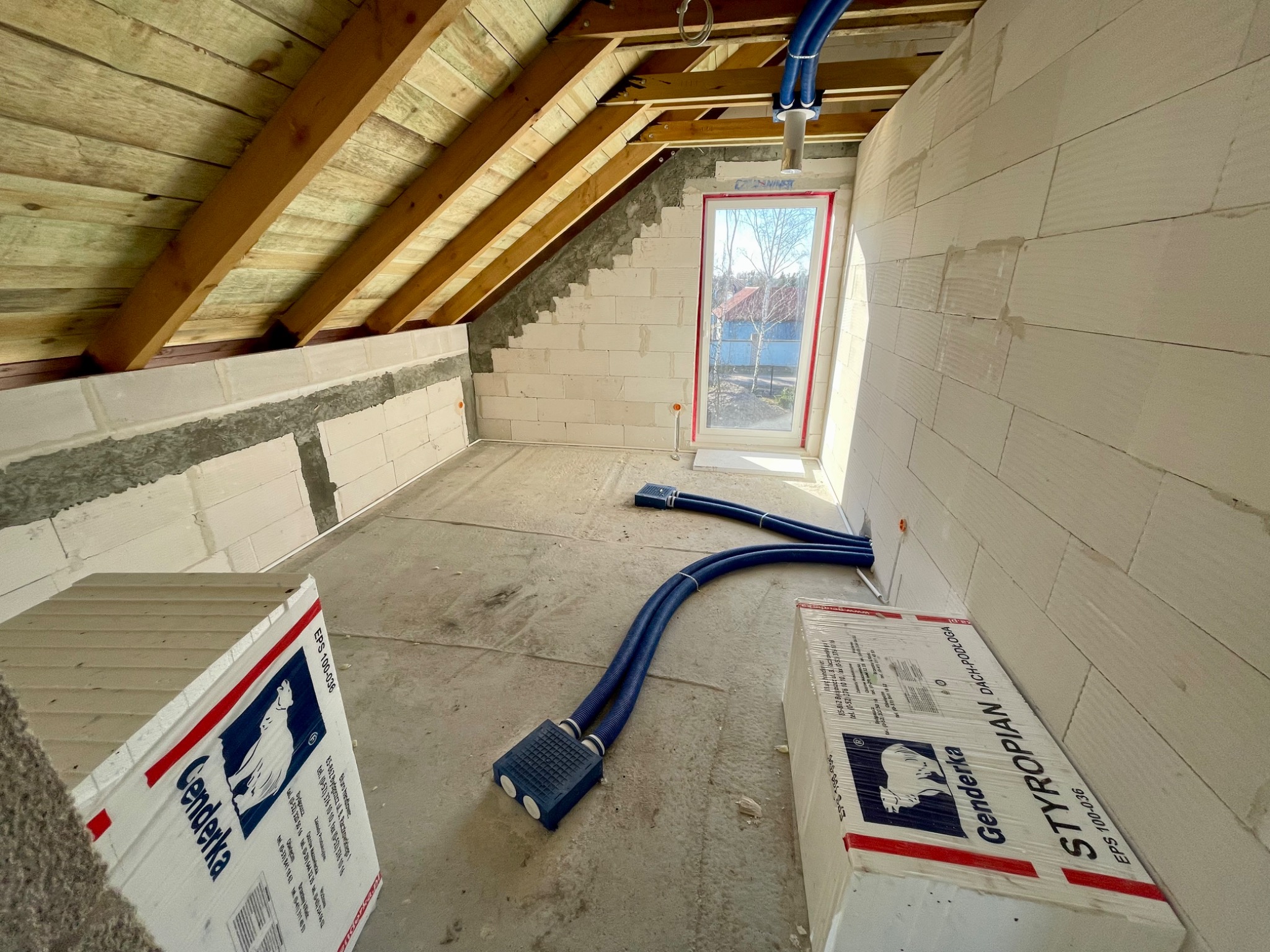 Poddasze w trakcie budowy z widocznymi elementami instalacji wentylacyjnej, w tym niebieskimi rurami i skrzynkami rozdzielczymi, oraz materiałami izolacyjnymi firmy Genderka.