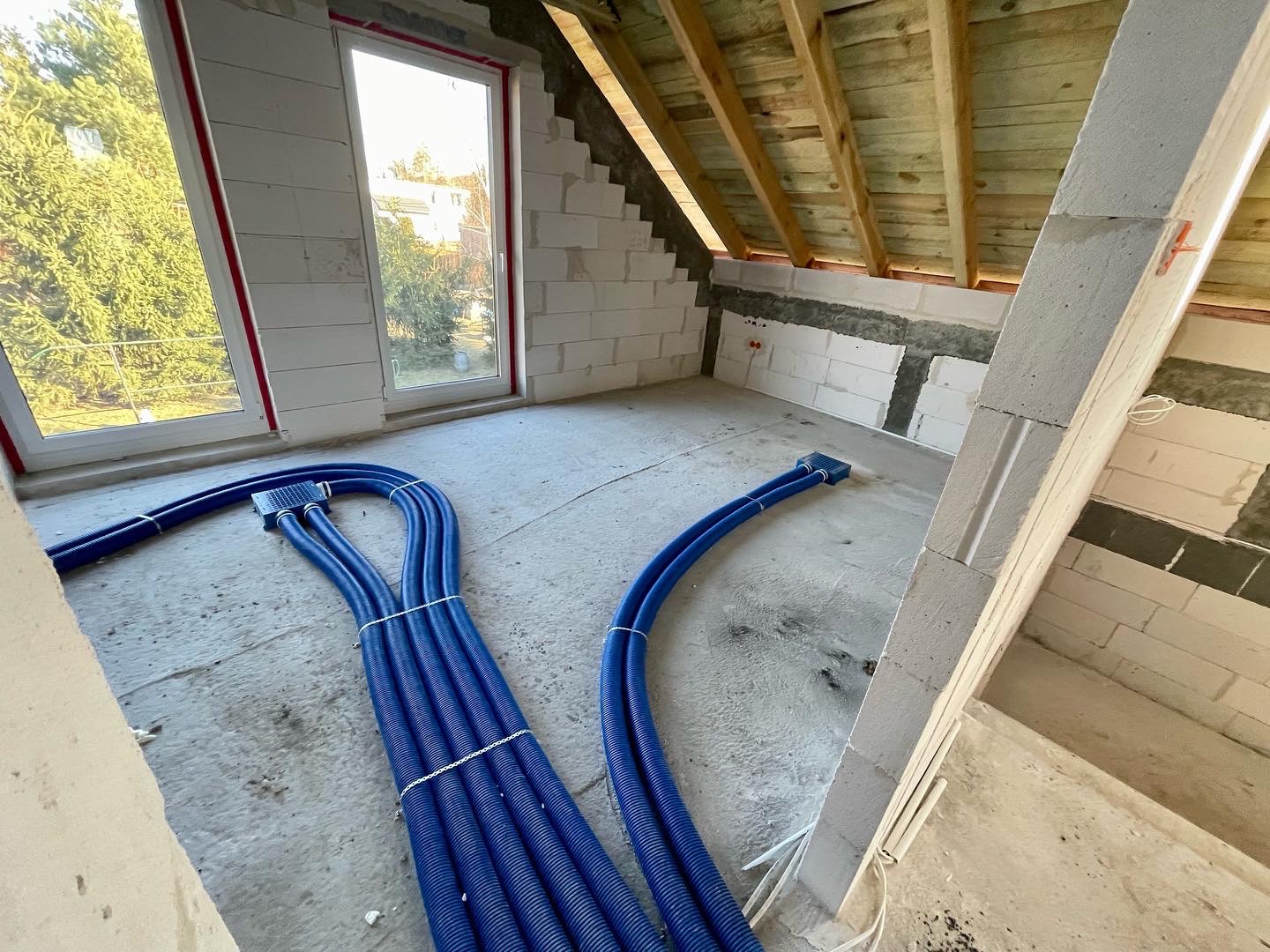 Wnętrze poddasza w trakcie prac wykończeniowych, widoczne niebieskie rury wentylacyjne na podłodze, białe ściany z betonu komórkowego i drewniana konstrukcja dachu.